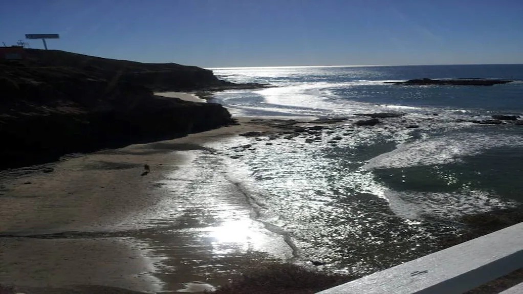 Sandy beach cove with gentle surf, rocky cliffs and bright sun reflecting on the water