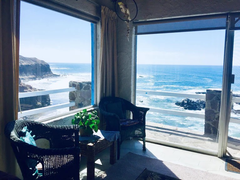 Wicker chairs and a potted plant in a corner sunroom with floor-to-ceiling windows overlooking the Pacific