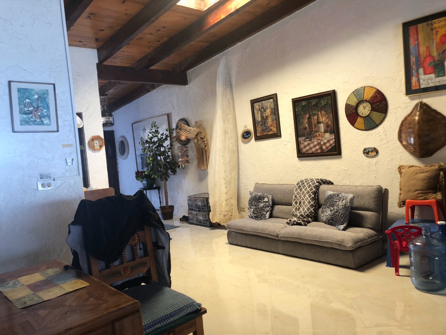 Open living and dining area with exposed wood-beam ceiling, white stucco walls hung with framed artwork, a sofa and a wooden dining table