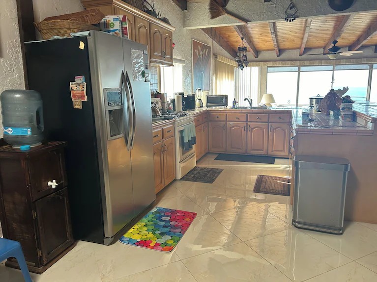 Kitchen with stainless steel refrigerator, wood cabinets, tiled counters and large windows looking out to the ocean